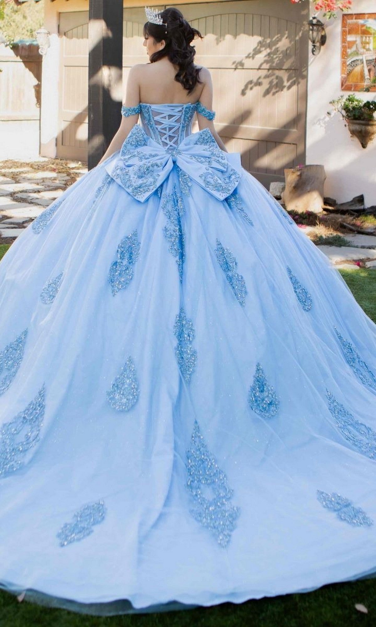 A woman stands outdoors facing away from the camera, wearing the Quinceanera Dress 8190J by Cinderella Couture—a blue ball gown featuring lace details and a large bow.